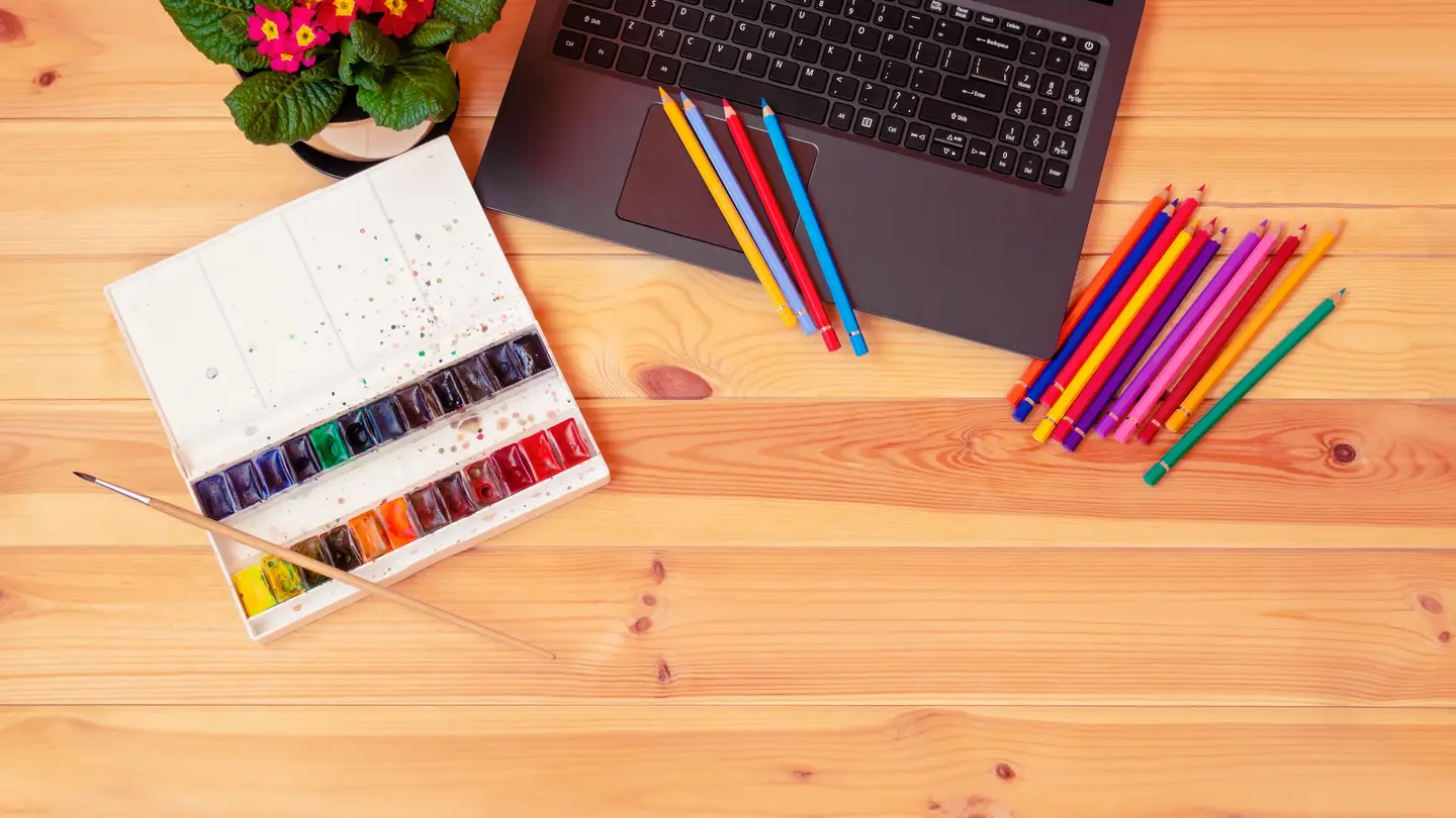 A watercolor paint set with a brush, colored pencils, a laptop and a potted primrose on a light wooden desk, seen from above
