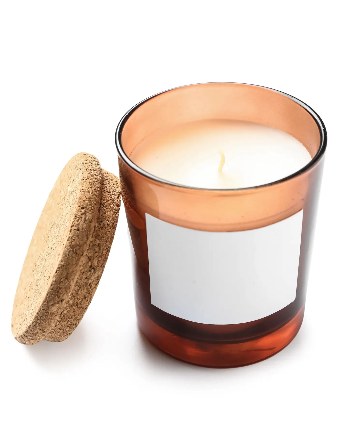 An amber glass candle jar with a white wax candle inside, a blank white label on the front and a cork lid leaning against it, on a plain white background