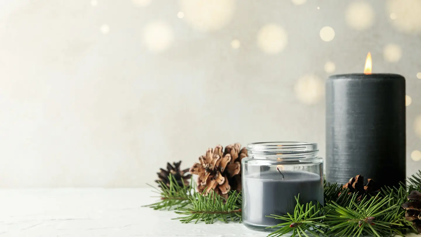 A glass jar candle and a lit dark pillar candle on a white table with pine branches and pinecones, soft lights glowing behind