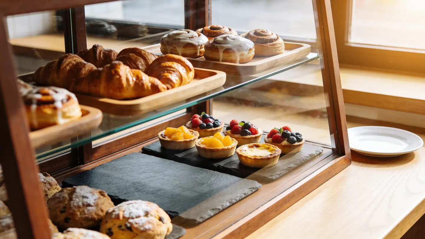 A wooden bakery display case with croissants, iced cinnamon rolls and fruit tarts in warm window light