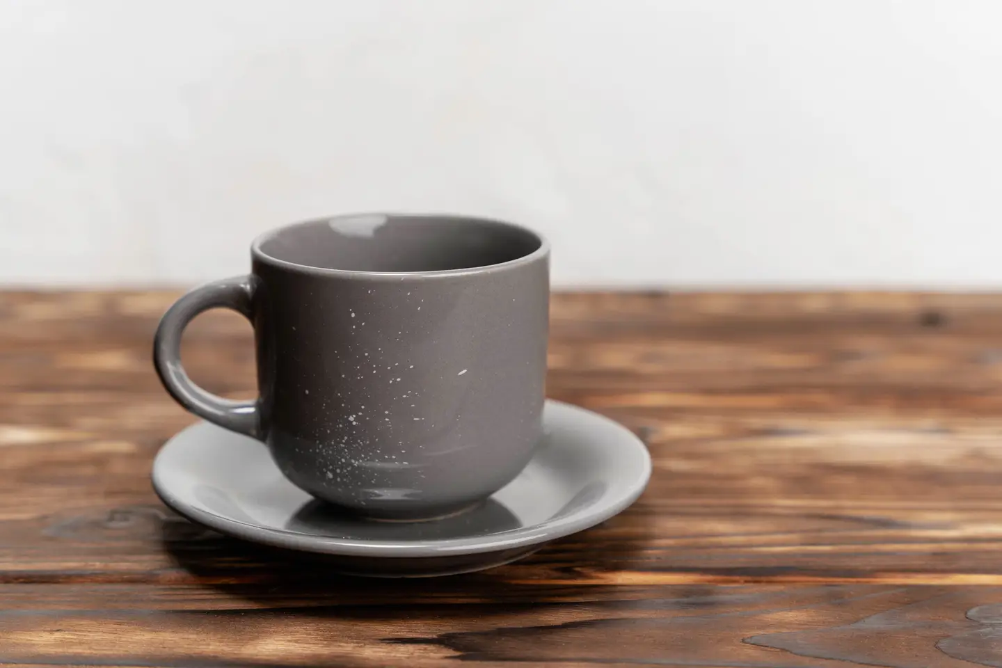 A gray speckled ceramic cup on a saucer on a dark wooden table, in front of a plain white wall