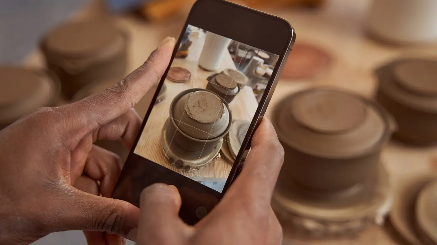 A potter holding a smartphone and photographing unfinished clay pots on a studio shelf