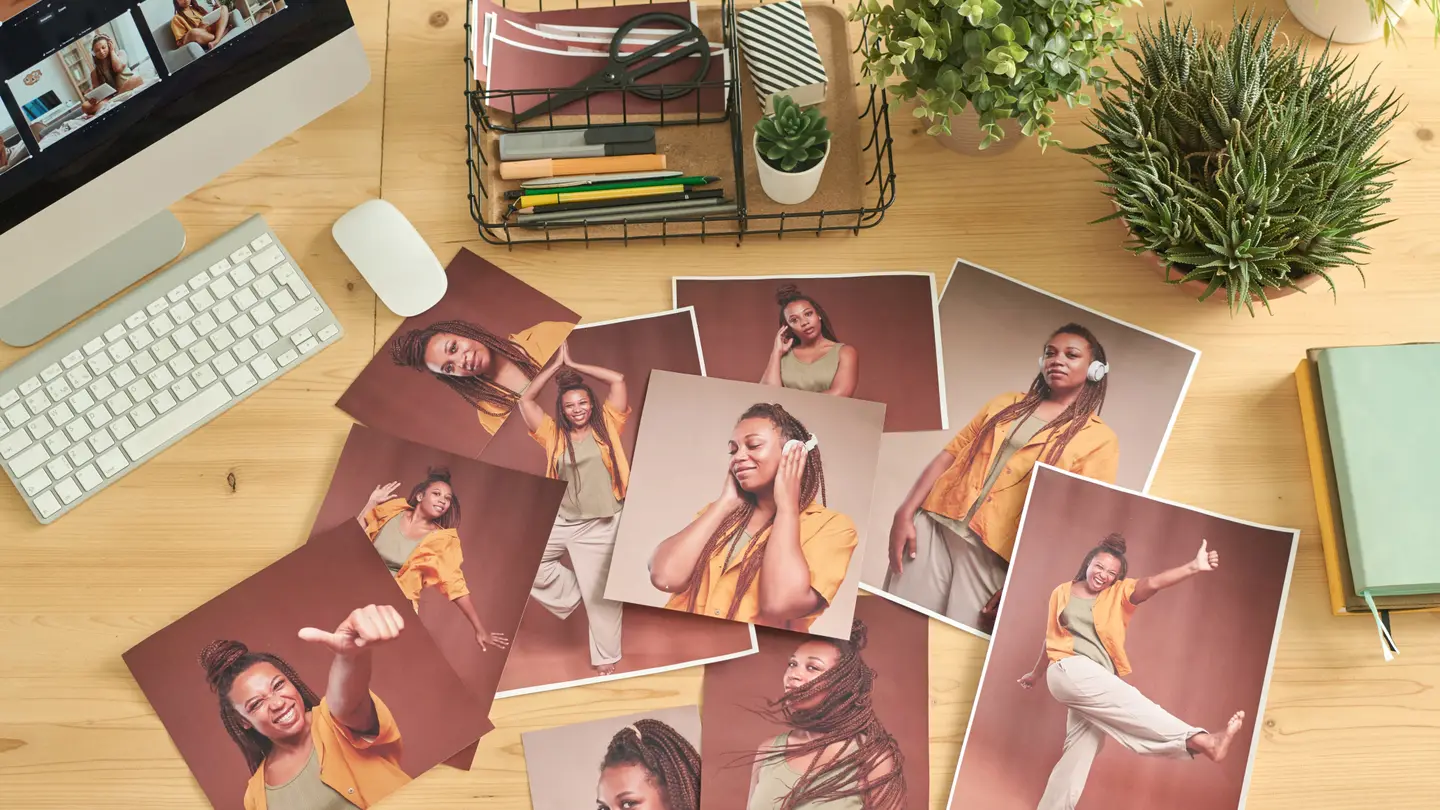 A wooden desk seen from above with a keyboard, plants and a dozen printed portraits laid out for review