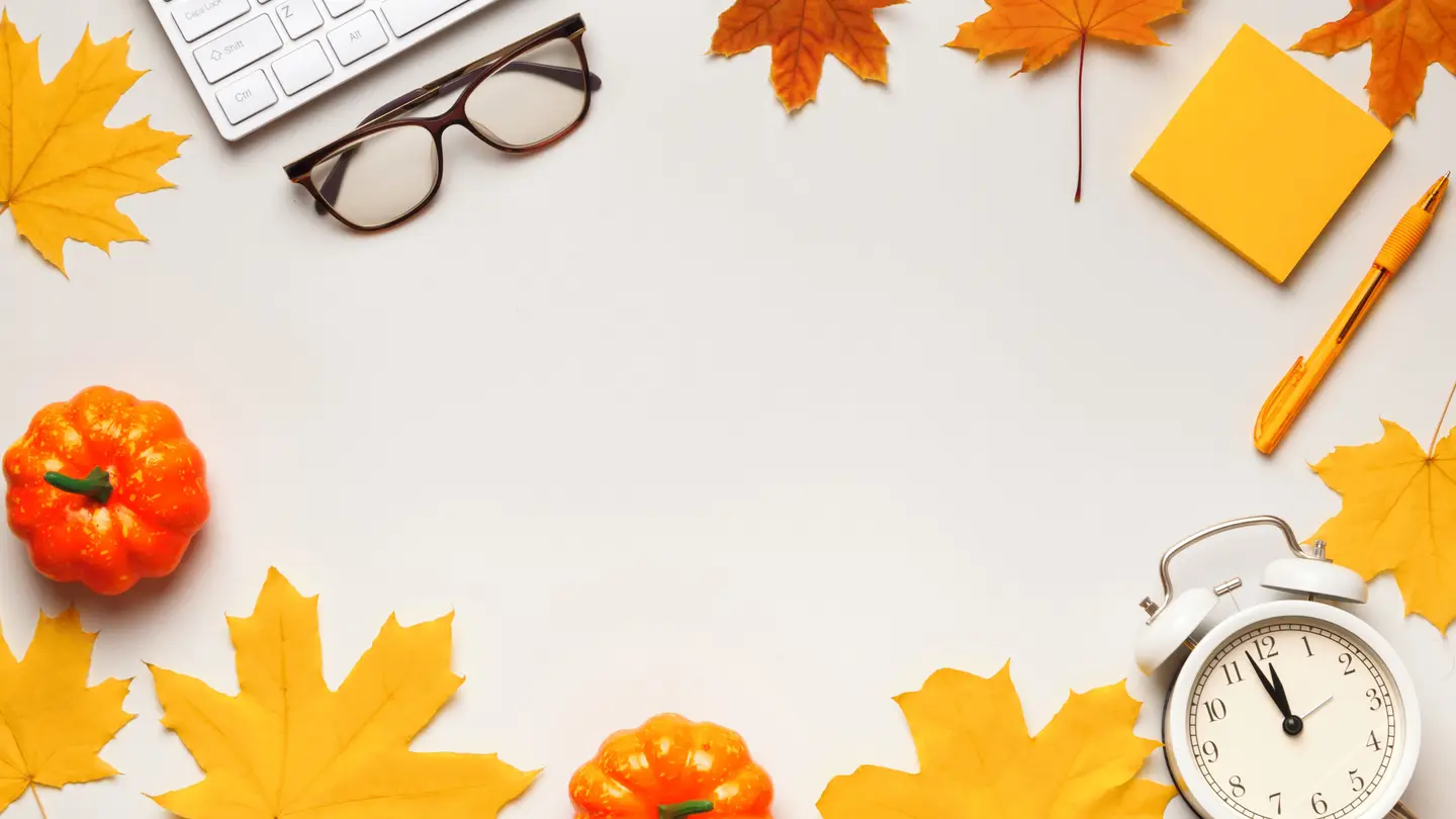 A flat lay of a white desk with a keyboard, glasses, an alarm clock, a yellow sticky note, small pumpkins and autumn maple leaves
