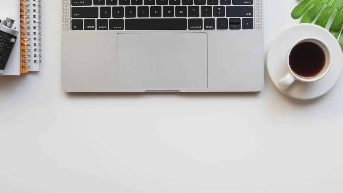 Top view of a white desk with a laptop, a camera, a notebook, a cup of coffee and a plant leaf