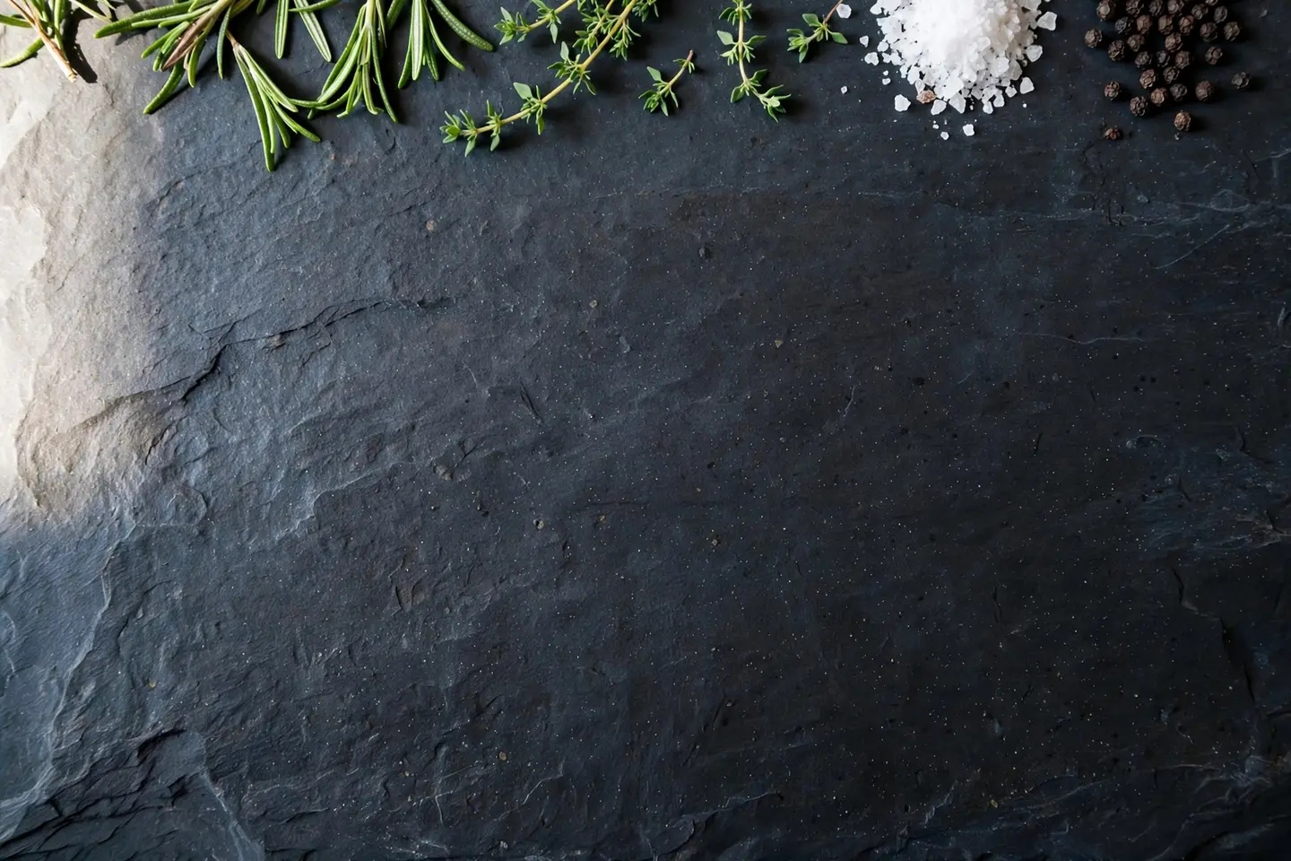 An AI image of a dark slate surface with rosemary, thyme, sea salt and peppercorns along the top edge and a large empty dark area below