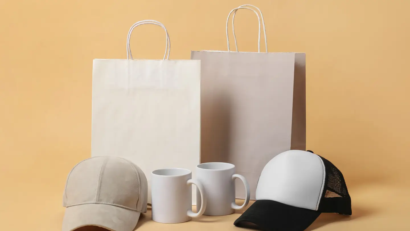 Blank merchandise on a beige background: two paper shopping bags, two white mugs and two caps ready for a design