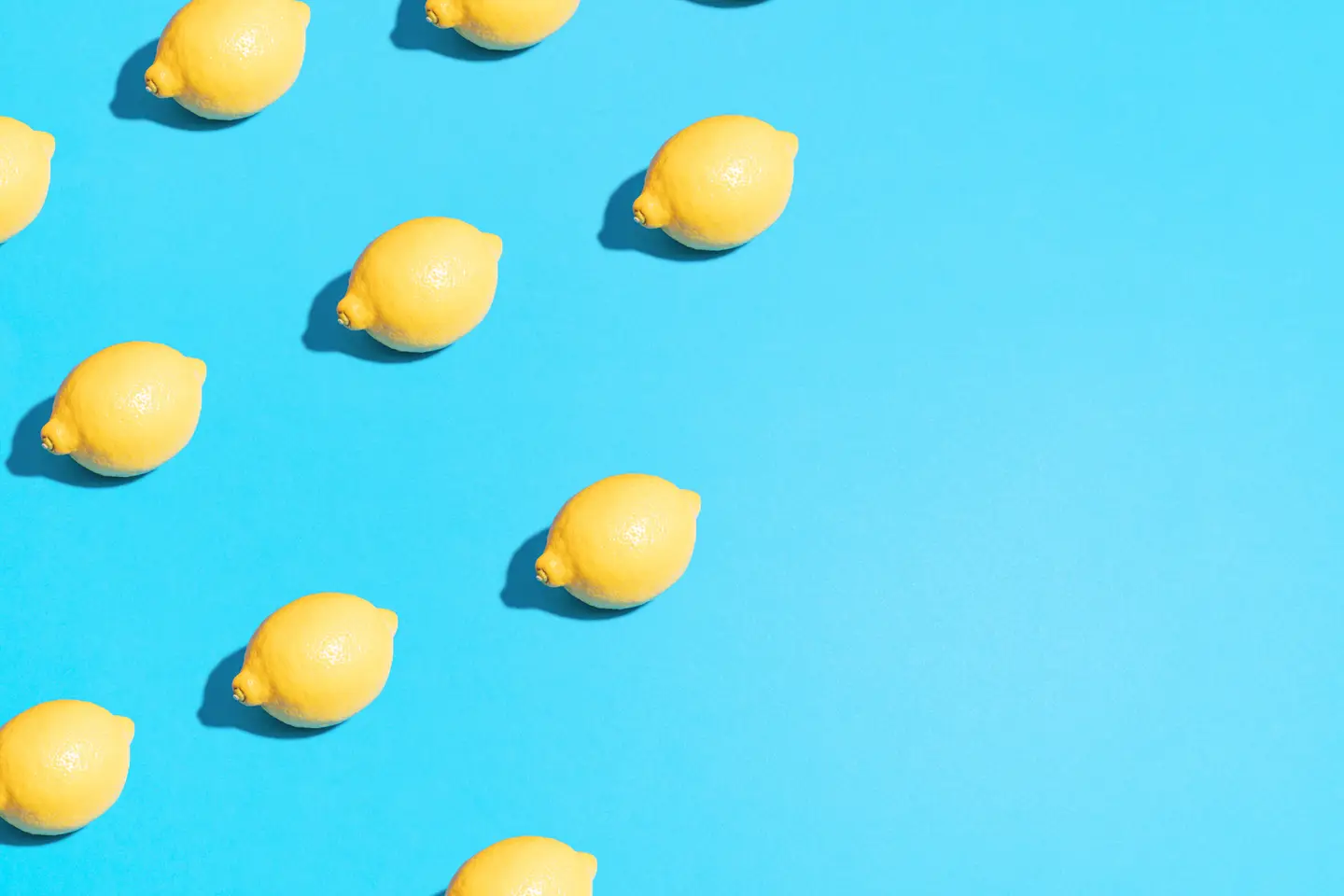 Rows of bright yellow lemons casting short shadows on a flat light blue background, with empty space on the right