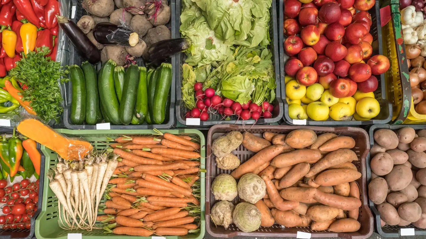Crates of colorful vegetables and fruit at a market stall seen from above: peppers, zucchini, lettuce, apples, carrots and potatoes