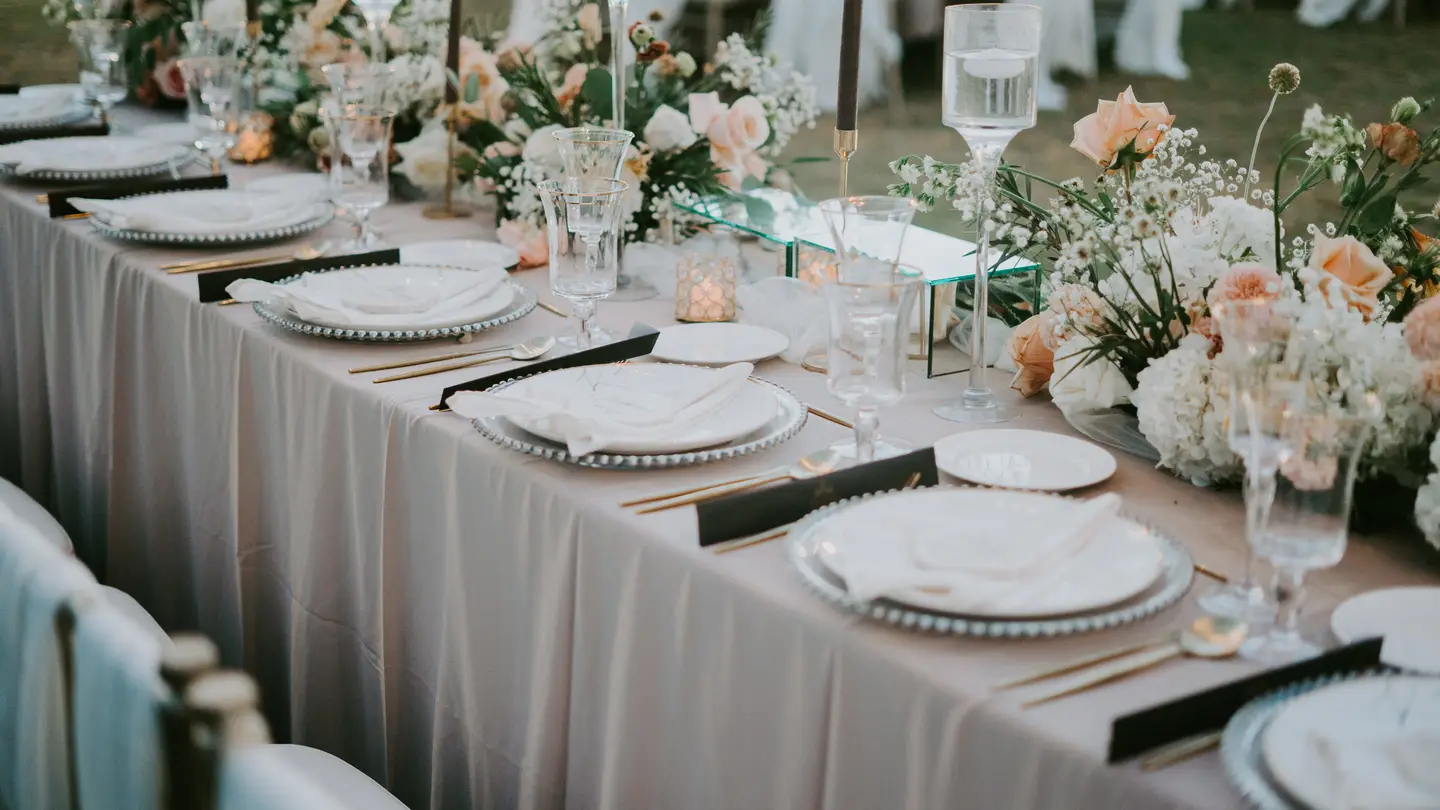 A long wedding reception table with a greige tablecloth, blush and peach flower arrangements, glassware, gold cutlery and black place cards